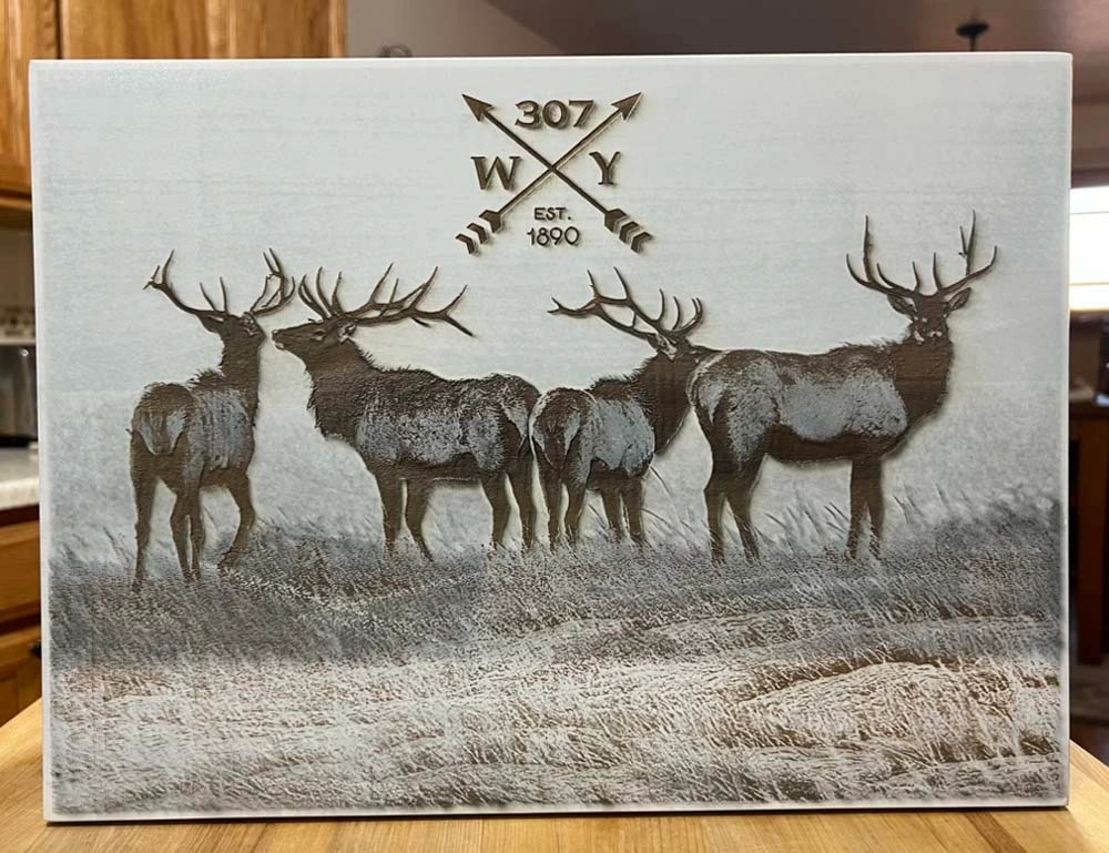 Engraved wood board showing a herd of elk with a '307 WY EST. 1890' crossed-arrows emblem, resting on a kitchen counter
