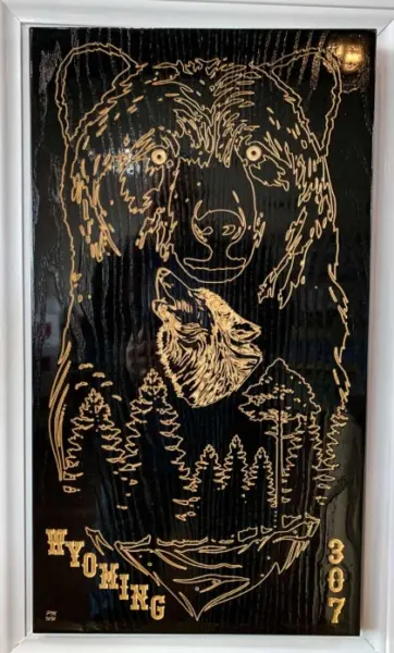 Laser-engraved artwork of a bear and howling wolf with pine trees, reading 'WYOMING 307,' displayed on black glass in a white frame